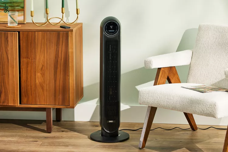 Un ventilador de torre negro y elegante, de pie sobre un suelo de madera en una sala de estar moderna. El ventilador tiene una pantalla digital en la parte superior y está situado entre un sillón de tela texturizada blanca y un aparador de madera. La imagen muestra cómo un ventilador de torre puede integrarse perfectamente como un electrodoméstico funcional y estilizado en la decoración del hogar.