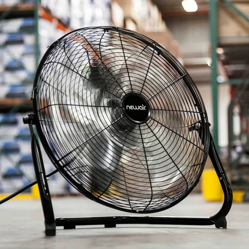 Un ventilador de suelo de alta velocidad, de metal y color negro, en funcionamiento dentro de un almacén. El ventilador está en un soporte tubular que le permite inclinarse y sus aspas de metal están borrosas por el movimiento, indicando que está encendido. La imagen captura un ventilador potente y robusto diseñado para una máxima circulación de aire en espacios amplios.