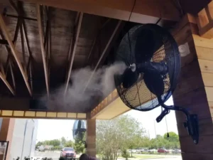 Un ventilador nebulizador de pared, de color negro, montado en el exterior de un patio cubierto con techo de madera. El ventilador está en pleno funcionamiento, rociando una fina nube de niebla de agua desde su centro para enfriar el aire. Esta imagen demuestra una solución de enfriamiento efectiva para espacios al aire libre como terrazas o restaurantes.