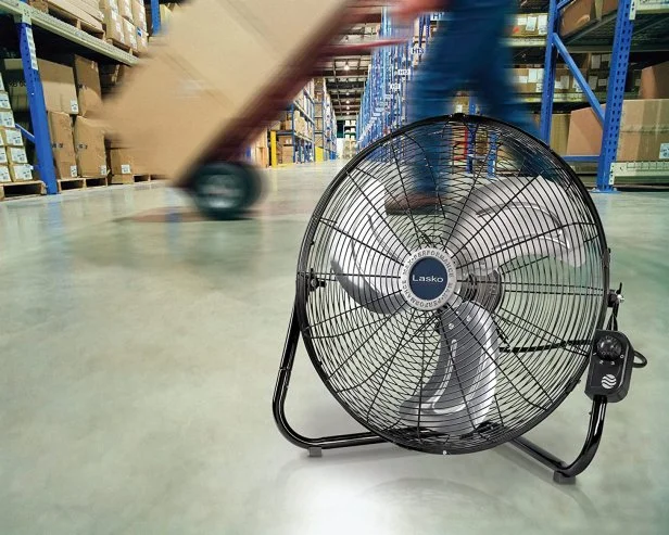 Un ventilador de suelo metálico de color negro con aspas plateadas, colocado en el suelo de hormigón de un almacén. El ventilador está enfocado en primer plano, mientras que el fondo muestra el movimiento borroso de un trabajador transportando cajas entre estanterías industriales. La imagen ilustra la potencia y la aplicación de estos ventiladores en entornos de trabajo logísticos y concurridos.