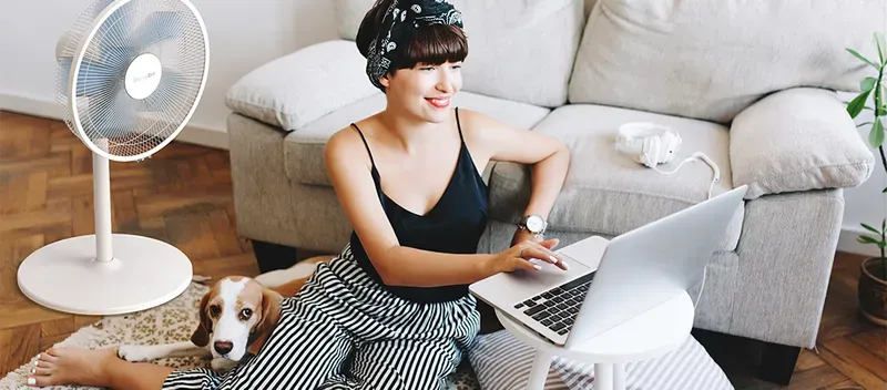 Una fotografía de estilo de vida que muestra a una mujer joven sentada en el suelo de su sala de estar, trabajando en un ordenador portátil, acompañada por su perro beagle. A su lado, un moderno ventilador de pedestal blanco está en funcionamiento, proporcionando una brisa fresca. La imagen captura un ambiente doméstico cómodo, relajado y fresco, ideal para el trabajo en casa o el descanso.